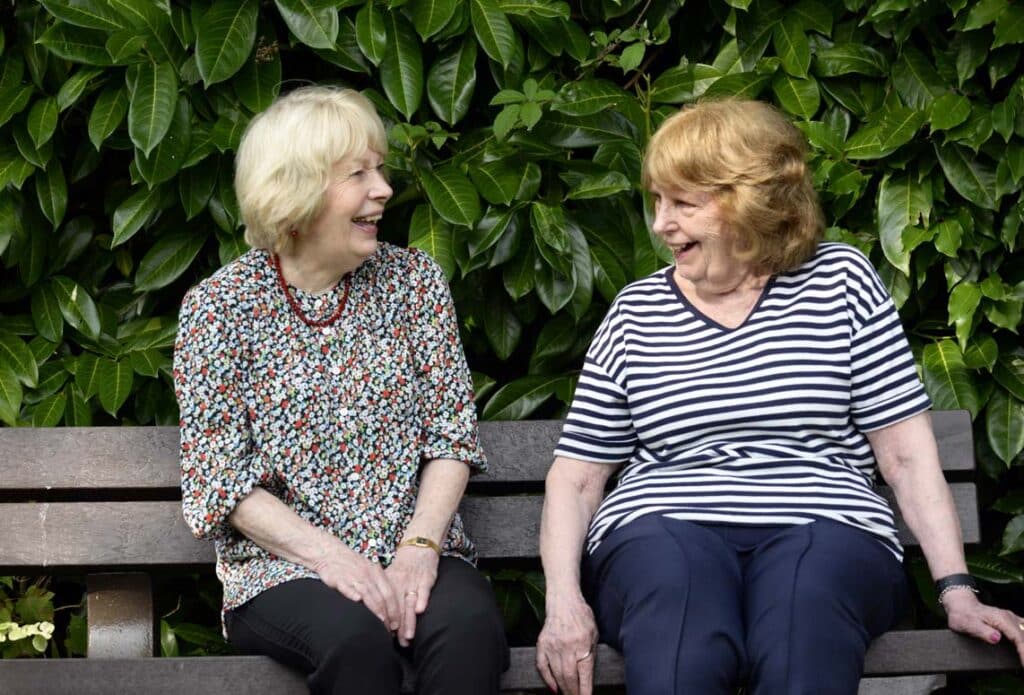 Two older women sitting outside on a garden bench talking and laughing