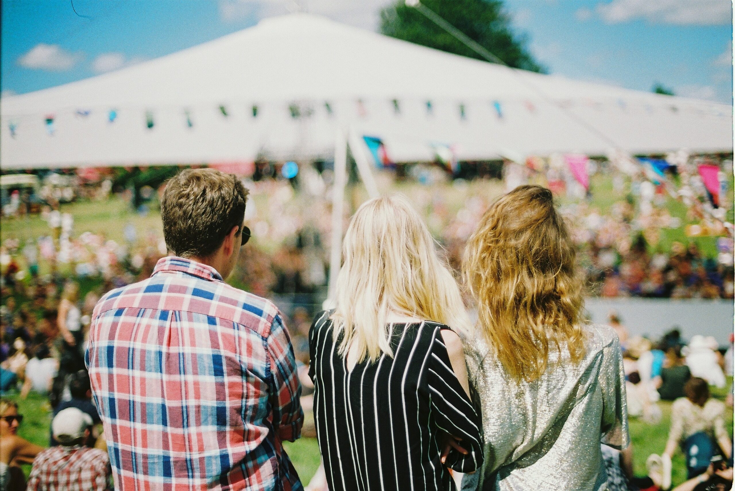 Backs of three people facing a crowd, a marquee and bunting on a sunny day